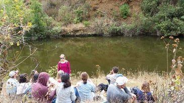 A woman engages a group of students in sustainability education on the bank of a river