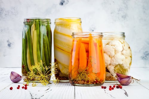 Four mason jars with pickled vegetables inside stand in a line in front of a nondescript background.